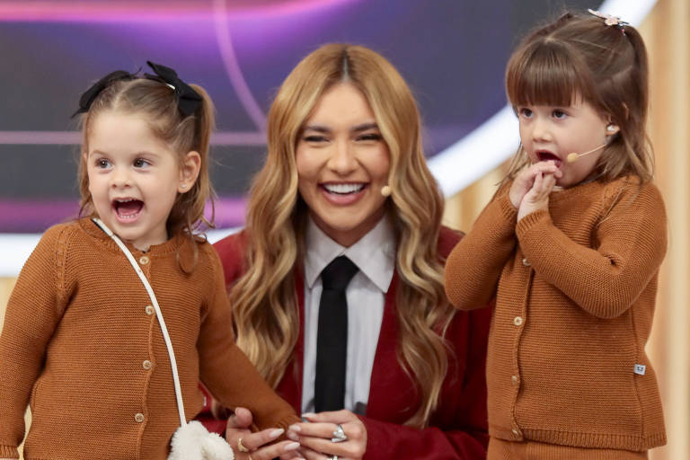 Uma mulher sorridente está agachada no centro da imagem, vestindo um vestido vermelho e uma camisa branca com gravata. Ao seu lado, duas meninas pequenas, também sorrindo, estão vestidas com roupas marrons. A cena parece ser de um programa de televisão, com um fundo colorido e decorativo.