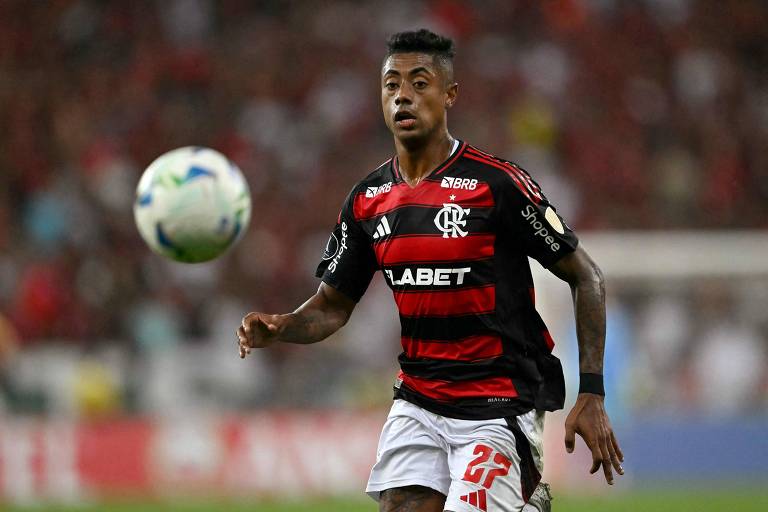 Um jogador de futebol está em campo, vestindo um uniforme listrado em vermelho e preto, representando o time Flamengo. Ele está correndo em direção à bola, que está flutuando no ar. O fundo mostra uma multidão de torcedores, com bandeiras e um ambiente de jogo vibrante.