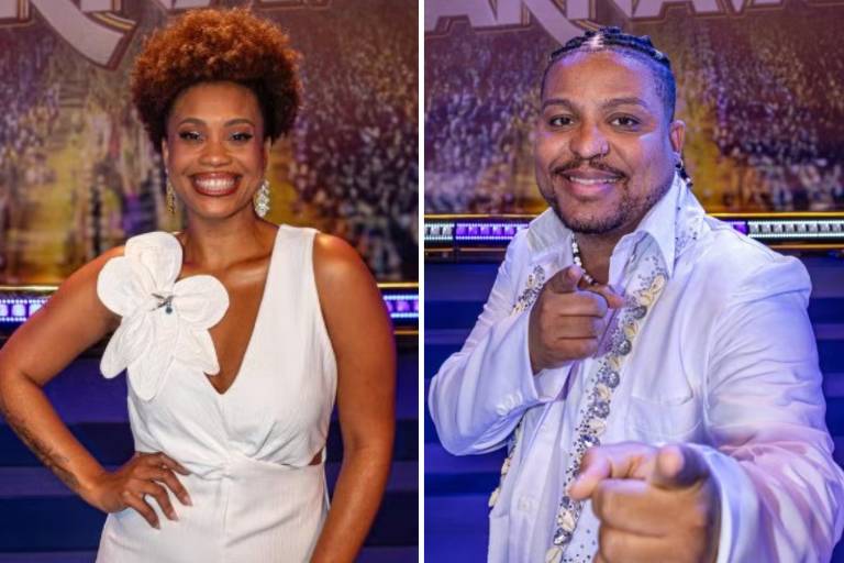 A imagem mostra duas pessoas em um cenário de programa de televisão. À esquerda, uma mulher com cabelo cacheado e volumoso, vestindo um vestido branco com uma flor grande na alça. À direita, um homem com cabelo trançado, usando uma camisa branca com detalhes brilhantes, apontando para a câmera. O fundo é decorado com elementos festivos relacionados ao carnaval.