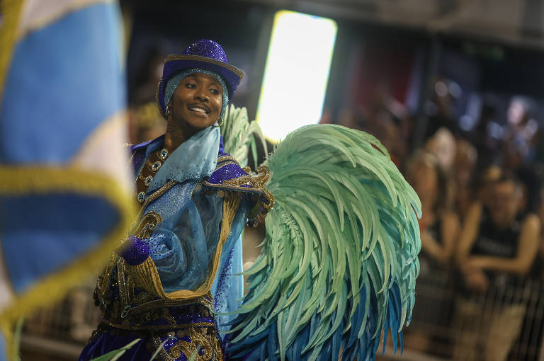 A imagem mostra um participante de um desfile de Carnaval, vestido com um traje colorido e brilhante. O traje é predominantemente azul, com detalhes em verde e dourado, e possui penas grandes que se assemelham a asas. O participante está sorrindo e posando para a câmera, enquanto ao fundo é possível ver uma multidão assistindo ao desfile.