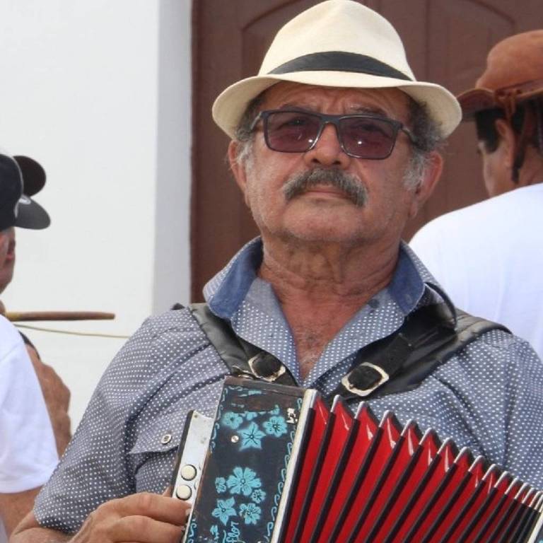 Um homem de óculos escuros e chapéu claro segura um acordeão. Ele está vestido com uma camisa cinza e parece estar em um ambiente ao ar livre, com outras pessoas ao fundo. O acordeão é vermelho e azul, e o homem tem uma expressão séria.