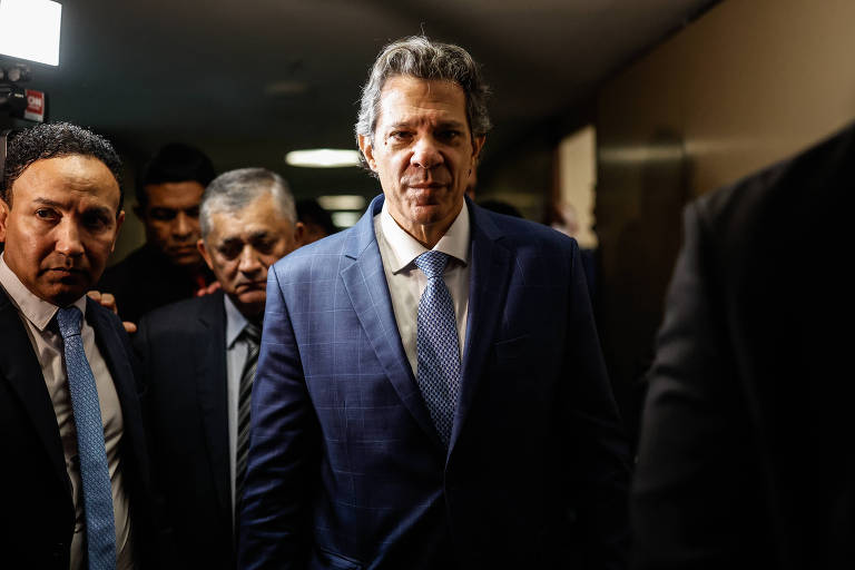 A imagem mostra um grupo de homens em um corredor. Um homem em destaque, com cabelo grisalho e terno azul, está caminhando em direção à câmera, enquanto outros homens o acompanham ao fundo. O ambiente parece ser interno, com iluminação artificial e paredes neutras.