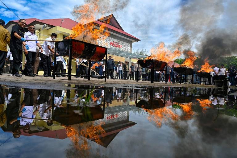 A imagem mostra um grupo de pessoas em um protesto, com várias barricas em chamas ao fundo. O céu está parcialmente nublado e há uma grande quantidade de fumaça saindo do fogo. No primeiro plano, é possível ver o reflexo das barricas em chamas na água acumulada no chão, criando um efeito visual interessante. As pessoas estão vestidas de maneira casual e algumas estão segurando objetos, possivelmente ferramentas ou bandeiras.