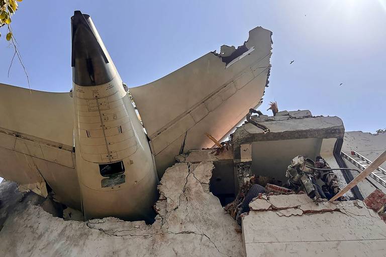 A imagem mostra a parte traseira de um avião colidido com um edifício. A fuselagem do avião está parcialmente inserida na estrutura do prédio, que apresenta danos significativos, com paredes quebradas e destroços visíveis. O céu está claro e ensolarado, e há alguns pássaros voando ao fundo.