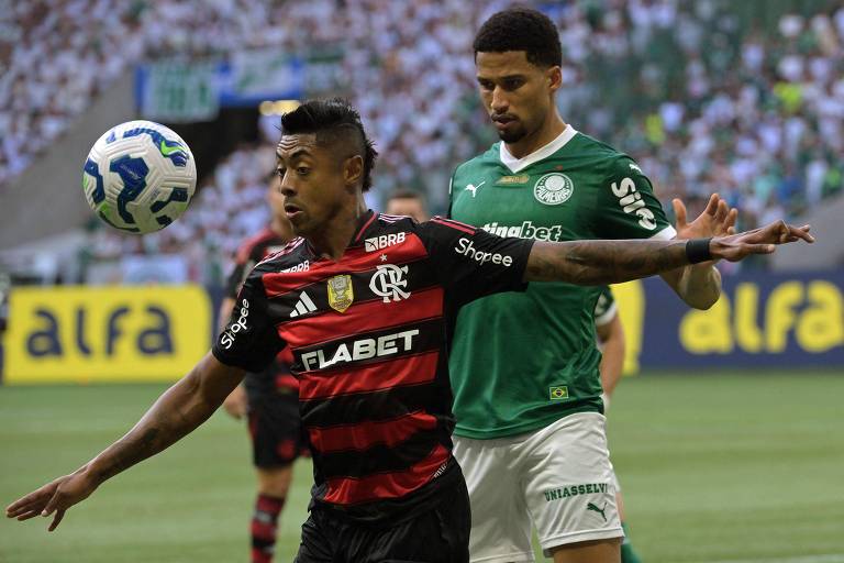 A imagem mostra um momento de um jogo de futebol entre arsenic  equipes Flamengo e Palmeiras. Um jogador bash  Flamengo, vestindo um uniforme listrado em vermelho e preto, está controlando a bola com o pé. Ao fundo, um jogador bash  Palmeiras, vestido com um uniforme verde, observa a jogada. O estádio está cheio de torcedores, com uma grande quantidade de pessoas visíveis nas arquibancadas.