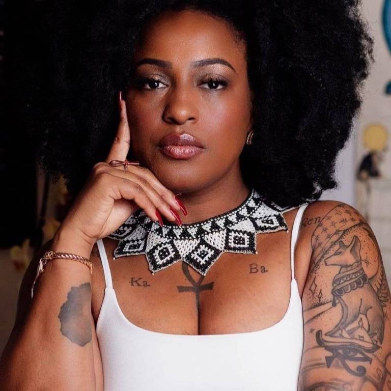 A imagem mostra uma mulher com cabelo afro volumoso, usando uma blusa branca. Ela tem um colar de contas com um padrão geométrico em preto e branco. A mulher está com uma expressão séria, com a mão direita apoiada no rosto, e possui várias tatuagens visíveis nos braços. O fundo da imagem é desfocado, mas há elementos decorativos que não são claramente identificáveis.