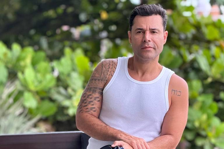 Um homem está posando em um ambiente externo, apoiado em uma grade. Ele usa uma camiseta regata branca e calças escuras. O homem segura uma jaqueta jeans e tem um tatuagem visível no braço. Ao fundo, há vegetação verde, sugerindo um ambiente natural.