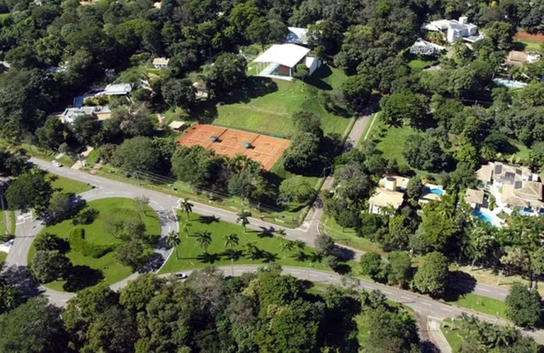 A imagem mostra uma vista aérea de uma área residencial cercada por vegetação. No centro, há um campo de tênis de terra batida. À direita, destaca-se uma casa moderna com um telhado branco. A área é bem arborizada, com várias árvores e plantas ao redor. Estradas circulares e retas estão visíveis, com alguns carros estacionados e em movimento. O ambiente é predominantemente verde, refletindo um espaço tranquilo e residencial.