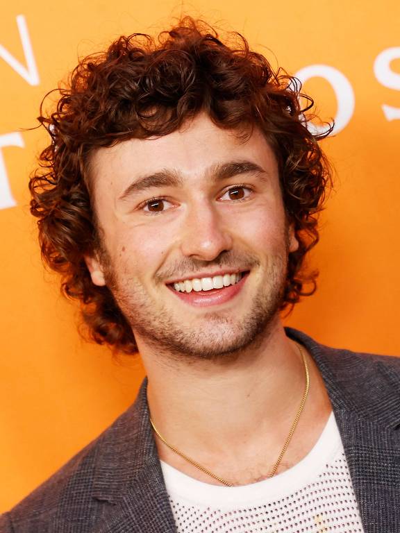 Um homem com cabelo cacheado e um sorriso largo está posando para a foto em frente a um fundo laranja. Ele usa uma jaqueta escura sobre uma camiseta branca com detalhes em malha. O fundo apresenta o texto 'AMAZON FILM STUDIOS' em letras brancas.