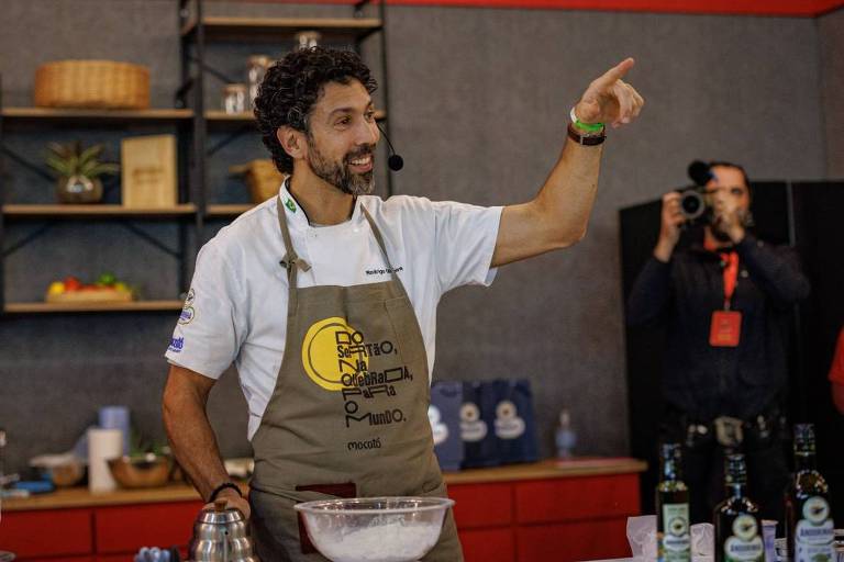 Um chef de cozinha, usando um avental, está em uma cozinha moderna, gesticulando enquanto fala. Ao seu lado, uma criança observa atentamente. Na mesa, há ingredientes e utensílios de cozinha, incluindo tigelas, garfos e garrafas de azeite. Ao fundo, um fotógrafo está registrando o momento, e uma mulher sorri, assistindo à demonstração.