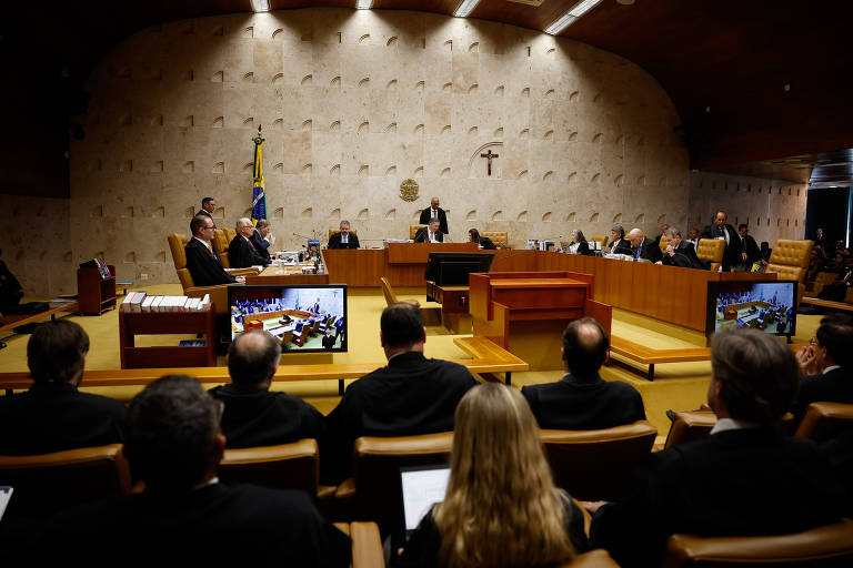 A imagem mostra uma sessão do Supremo Tribunal Federal do Brasil. No centro, há uma mesa com vários juízes e um presidente da sessão, todos em trajes formais. Ao fundo, há uma bandeira do Brasil e um crucifixo na parede. Na frente, há uma plateia com pessoas sentadas, algumas delas com trajes escuros. Em duas telas de projeção, é possível ver informações relacionadas à sessão.