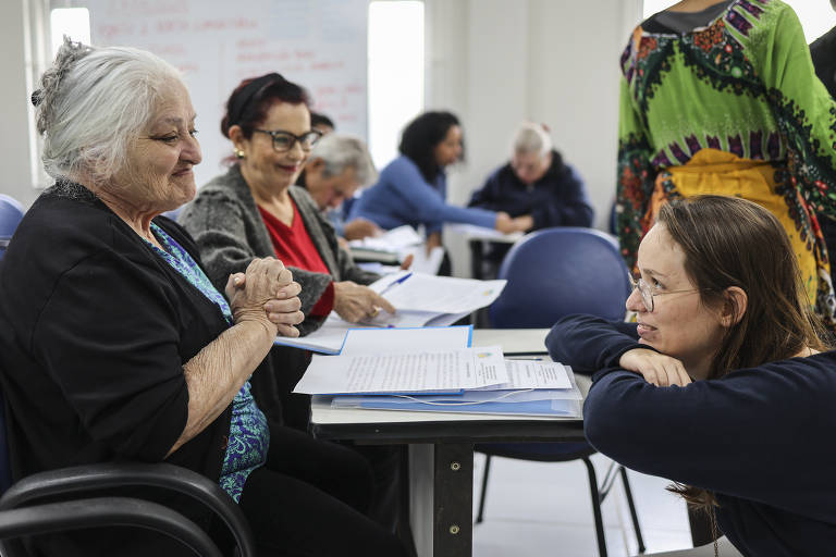 A imagem mostra um grupo de pessoas em um ambiente de sala de aula ou reunião. Duas mulheres estão sentadas à frente, uma com cabelo grisalho e a outra com cabelo castanho claro, ambas parecendo envolvidas em uma conversa. Ao fundo, outras pessoas estão sentadas e interagindo. A mulher de cabelo grisalho está com as mãos juntas, enquanto a outra mulher está apoiando a cabeça nas mãos, olhando atentamente. O ambiente é iluminado e as pessoas estão vestindo roupas casuais.