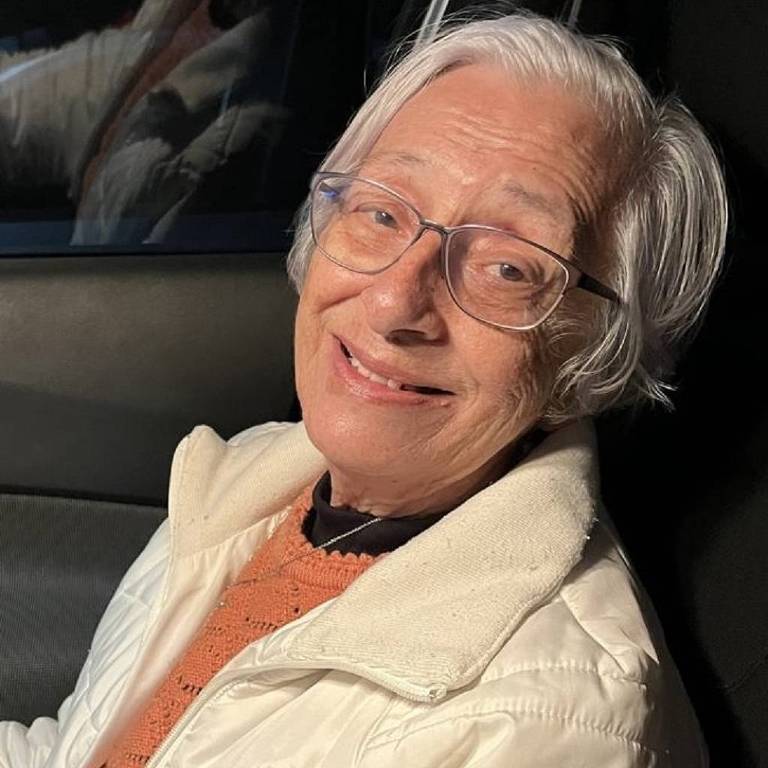 Uma mulher idosa com cabelo branco e óculos, sorrindo enquanto está sentada em um carro. Ela usa um casaco claro e uma blusa laranja por baixo. O fundo é um interior de carro, com reflexos visíveis.