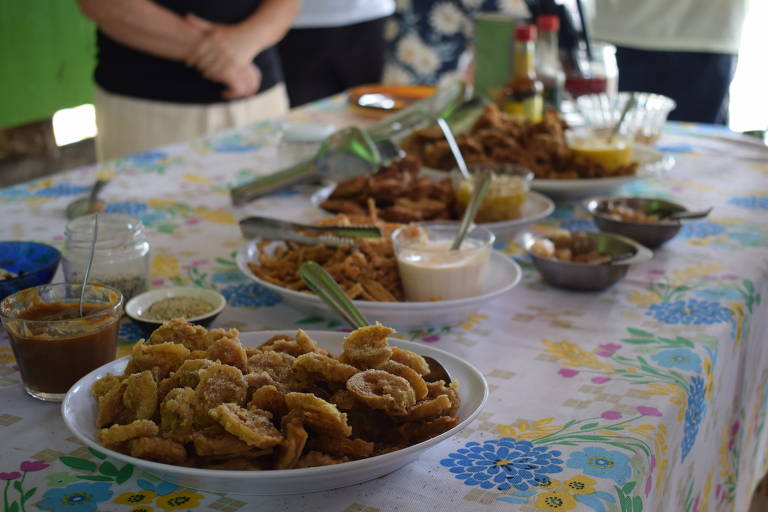 A imagem mostra uma mesa com diversos pratos de comida típica. Há várias tigelas e pratos com diferentes tipos de alimentos, incluindo frituras e molhos. O fundo é decorado com uma toalha de mesa colorida e há pessoas parcialmente visíveis ao fundo, mas não é possível identificar detalhes sobre elas.