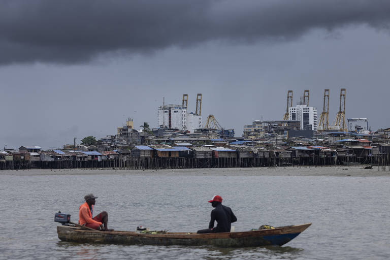 A imagem mostra dois homens em um pequeno barco de madeira, pescando em uma área aquática. Ao fundo, há uma cidade com edifícios e estruturas industriais, com um céu nublado e escuro. A água parece calma, e as casas na margem estão construídas sobre palafitas.