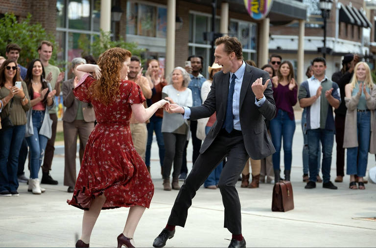 Uma cena de dança em um espaço público, onde um homem e uma mulher estão dançando juntos. A mulher usa um vestido vermelho com estampas florais e o homem está vestido com um terno escuro e uma camisa clara. Eles estão cercados por uma multidão de pessoas que assistem e aplaudem. O ambiente parece ser uma área comercial ao ar livre, com lojas ao fundo e um clima alegre.