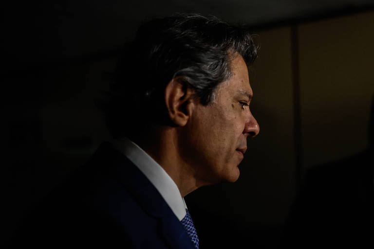 A imagem mostra o perfil de um homem com cabelo escuro e levemente ondulado, usando um terno escuro e uma camisa branca. A iluminação é baixa, destacando o contorno do rosto e criando um efeito de sombra. O fundo é desfocado, sugerindo um ambiente interno.