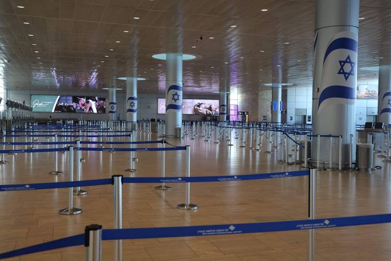 A imagem mostra uma área de check-in de um aeroporto, com várias filas organizadas por cordas azuis. O espaço é amplo e possui colunas com a bandeira de Israel. Ao fundo, há telas de exibição e o piso é de madeira clara. O ambiente parece estar vazio, sem pessoas visíveis.