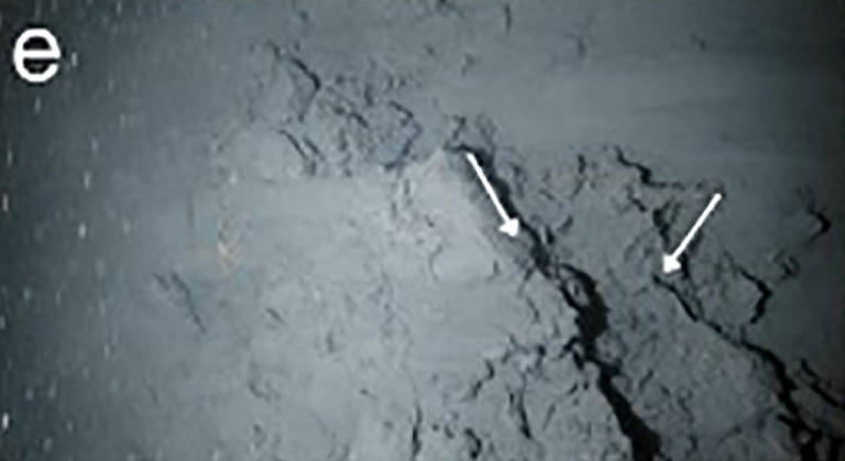 A imagem mostra uma superfície do fundo do mar, com uma textura irregular e rachaduras visíveis. Duas setas brancas apontam para as rachaduras, destacando-as. O fundo é predominantemente escuro, com uma aparência de sedimento ou rocha.