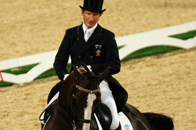 Um cavaleiro vestido com um traje formal, incluindo um chapéu alto, está montado em um cavalo preto durante uma competição de adestramento. O ambiente é uma arena coberta com piso de areia, e ao fundo, há uma faixa verde visível. O cavalo parece estar em movimento, e o cavaleiro mantém uma postura ereta e concentrada.