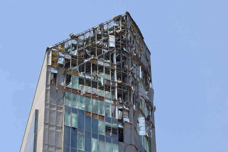 A imagem mostra a parte superior de um edifício com danos visíveis. A estrutura parece ter sofrido um colapso parcial, com janelas quebradas e partes da fachada expostas. O céu ao fundo é claro e azul, contrastando com a aparência danificada do edifício.