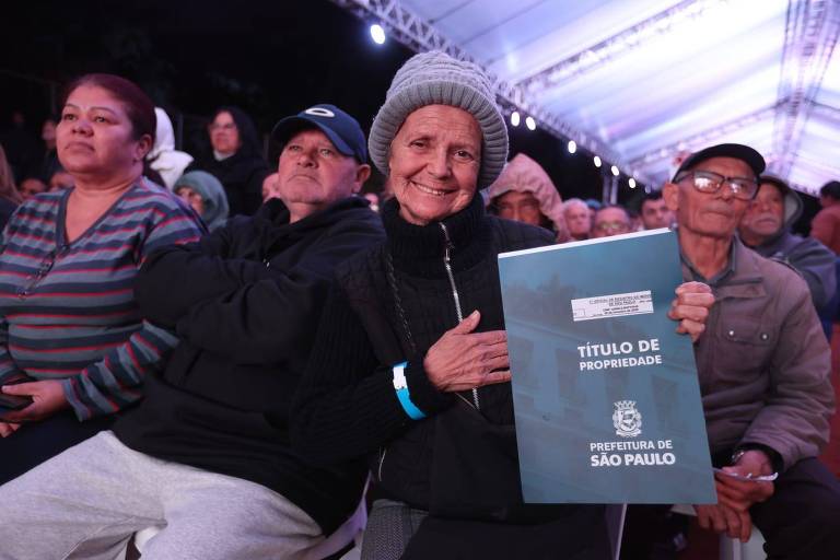 A imagem mostra um grupo de pessoas sentadas em um evento ao ar livre, sob uma estrutura coberta. No centro, uma mulher idosa sorri enquanto segura um documento azul intitulado 'TÍTULO DE PROPRIEDADE', que é emitido pela Prefeitura de São Paulo. Ao fundo, outras pessoas assistem ao evento, algumas com expressões de expectativa.