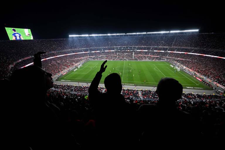 A imagem mostra um estádio de futebol lotado, com torcedores visíveis em primeiro plano, levantando os braços em celebração. O campo está bem iluminado e há uma tela grande exibindo uma partida. O ambiente é noturno, com muitas luzes acesas no estádio e uma atmosfera vibrante.