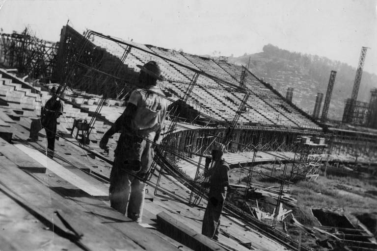 A imagem mostra um canteiro de obras com trabalhadores em atividade. No primeiro plano, três homens estão em uma estrutura de madeira, aparentemente em uma fase de construção. Ao fundo, há uma grande estrutura metálica em construção, com várias colunas e suportes visíveis. A paisagem ao fundo é montanhosa, e o céu parece claro. A foto é em preto e branco, o que dá um aspecto nostálgico à cena.