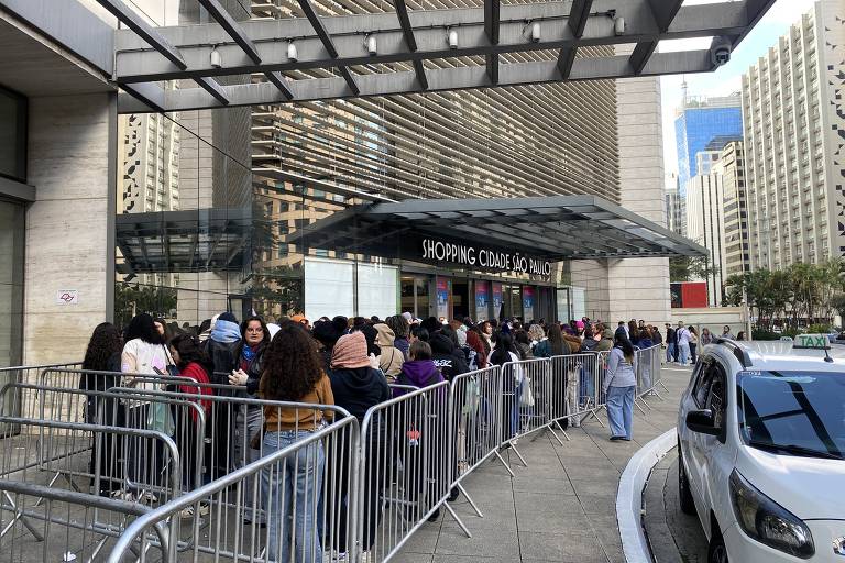 A imagem mostra uma longa fila de pessoas do lado de fora de um shopping. As pessoas estão organizadas em uma fila delimitada por grades de metal. O shopping é identificado como 'SHOPPING CIDAD JAGUARÉ'. Ao fundo, há prédios altos e modernos, e o céu está claro. Um carro branco está estacionado próximo à entrada do shopping.