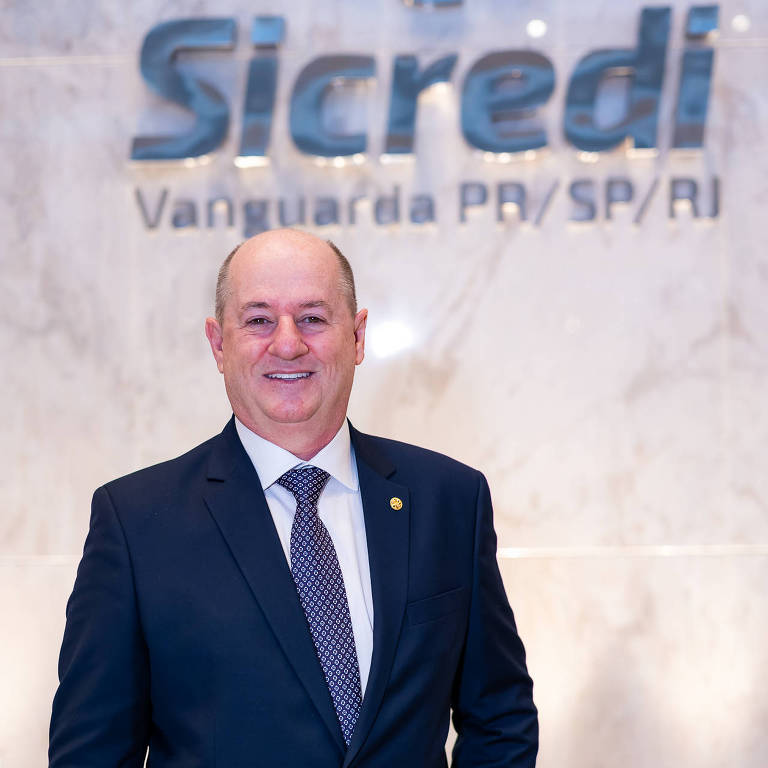 Um homem sorridente está posicionado em frente a um fundo com o logo da instituição financeira Sicredi. Ele usa um terno escuro e uma gravata, e parece estar em um ambiente de escritório bem iluminado. O logo do Sicredi é visível atrás dele, com a inscrição 'Vanguarda PD/SP/RJ'.