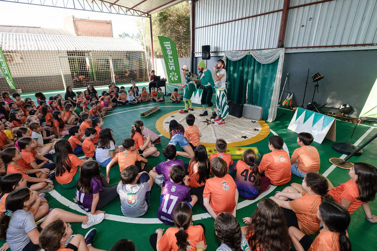 A imagem mostra um evento em um espaço coberto onde várias crianças estão sentadas em um círculo, assistindo a uma apresentação. As crianças estão vestidas com camisetas coloridas, principalmente em tons de laranja e algumas em outras cores. No centro, duas pessoas estão se apresentando, uma delas vestida com roupas brancas e verdes. Ao fundo, há banners verdes com a marca 'siredi' e equipamentos de som. O ambiente parece festivo e animado.