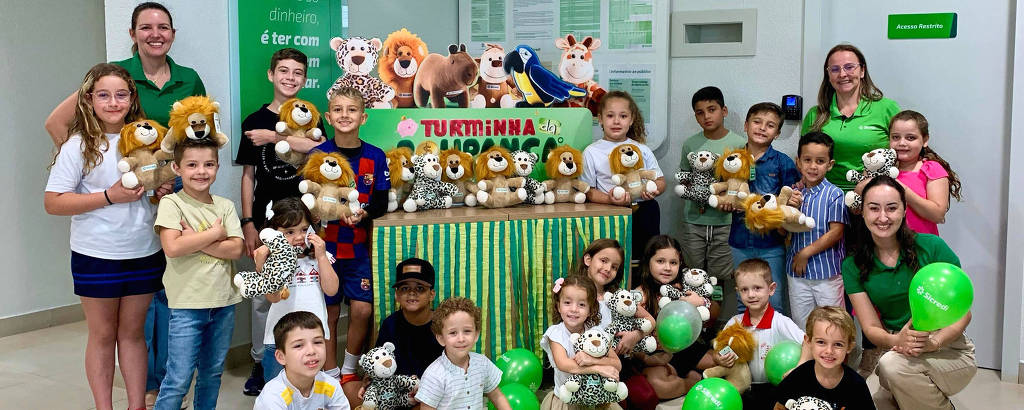 A imagem mostra um grupo de crianças e adultos em um ambiente interno, possivelmente uma sala de atividades. As crianças estão segurando pelúcias de animais e algumas têm balões verdes. Ao fundo, há uma mesa decorada com mais pelúcias e um banner que diz 'TURMINHA'. O ambiente é iluminado e as pessoas estão sorrindo, demonstrando alegria.