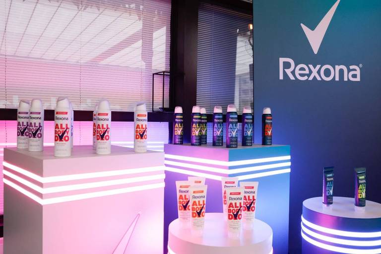 A imagem mostra uma exposição de produtos da marca Rexona, incluindo desodorantes em spray e em creme. Os produtos estão dispostos em suportes brancos iluminados com luzes coloridas, predominantemente em tons de rosa e azul. Ao fundo, há um painel com o logotipo da Rexona, que é um símbolo de verificação. A iluminação e a disposição dos produtos criam um ambiente moderno e atrativo.