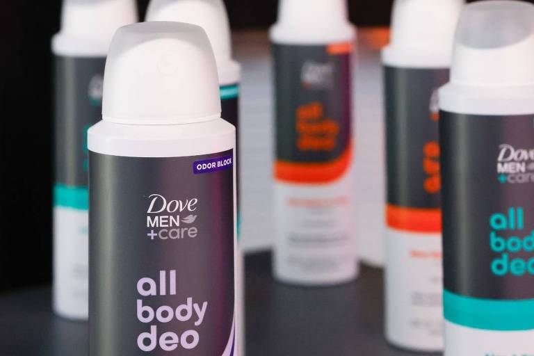 A imagem mostra várias latas de desodorante da marca Dove Men+Care em um fundo escuro. Em destaque, está uma lata de desodorante com a etiqueta 'all body deo' e a fragrância 'Fig & Suede'. As outras latas têm diferentes cores e rótulos, mas não são legíveis. O ambiente parece ser uma prateleira ou exibição de produtos.