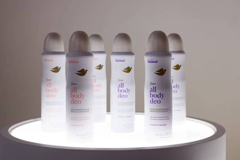 A imagem mostra vários frascos de desodorante da marca Dove dispostos em um suporte circular iluminado. Os frascos são brancos com tampas claras e apresentam a inscrição 'Dove all body deo' em diferentes cores, indicando diferentes fragrâncias. O fundo é neutro e destaca os produtos.