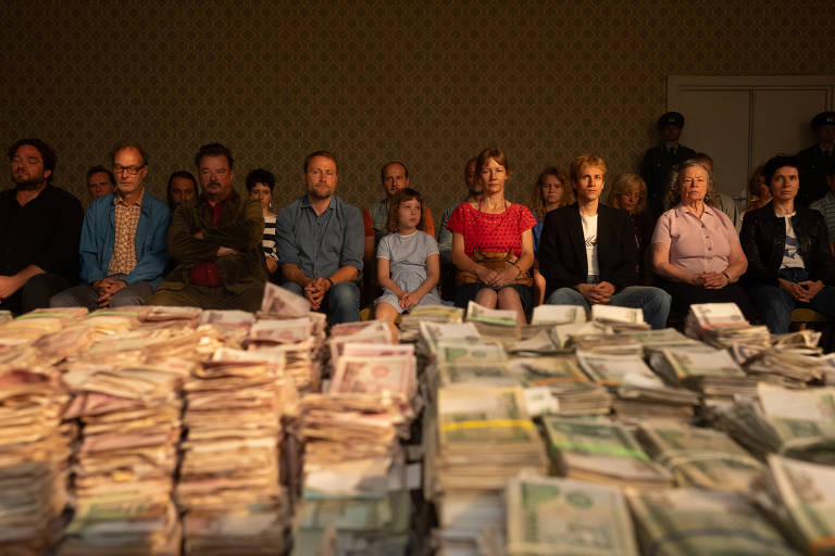 A imagem mostra um grupo de pessoas sentadas em uma sala, com uma grande quantidade de dinheiro em notas empilhadas à frente delas. As pessoas estão em uma disposição ordenada, com expressões neutras. O ambiente parece ser um espaço fechado, com paredes decoradas. A iluminação é suave, destacando o dinheiro em primeiro plano.