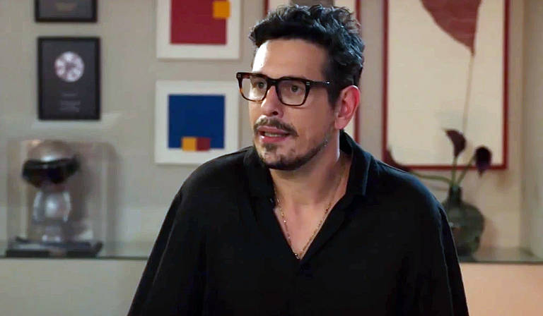 Um homem com cabelo escuro e barba, usando óculos, está em um ambiente interno. Ele veste uma camisa preta e parece estar em uma conversa, com uma expressão de atenção. Ao fundo, há quadros coloridos e um objeto decorativo em uma prateleira.