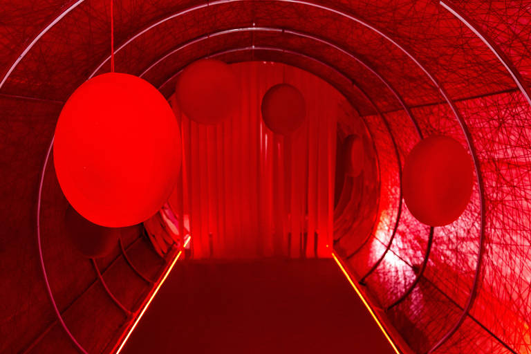 A imagem mostra um túnel com iluminação vermelha, decorado com esferas grandes e redondas penduradas no teto. As paredes do túnel são revestidas com uma malha que também é vermelha, criando um ambiente imersivo e vibrante. O chão é plano e há uma faixa de luz ao longo do caminho, destacando a entrada do túnel.