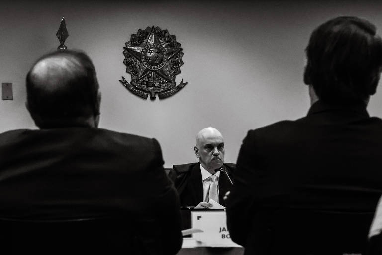 A imagem mostra uma reunião em um ambiente formal, com várias pessoas sentadas de costas. No centro, um homem de cabelo curto e calvo está em destaque, enquanto outros dois homens, um à esquerda e outro à direita, estão parcialmente visíveis. Ao fundo, há um emblema ou brasão na parede. A imagem é em preto e branco, conferindo um tom sério à cena.