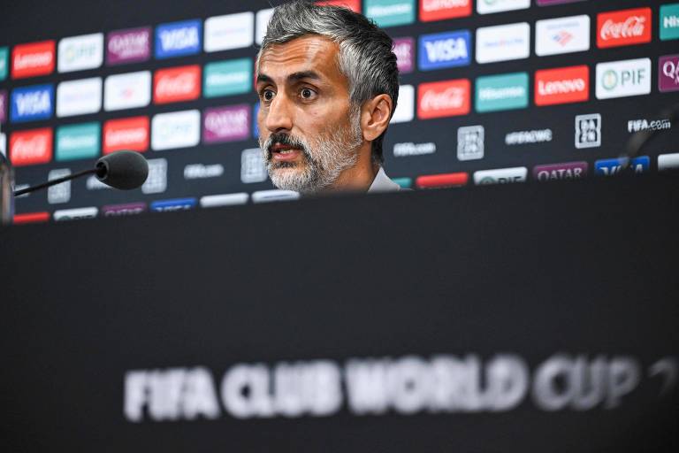 Um homem com cabelo grisalho e barba está sentado em uma mesa de coletiva de imprensa. Ele parece estar falando ou respondendo a perguntas. Ao fundo, há um painel com logotipos de patrocinadores e a frase 'FIFA CLUB WORLD CUP' visível na parte inferior da imagem.