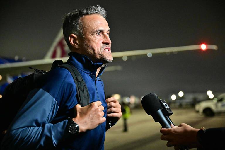Um homem com cabelo grisalho e uma expressão séria está em um aeroporto à noite. Ele usa uma jaqueta azul e carrega uma mochila nas costas. À sua frente, um repórter está segurando um microfone, aparentemente fazendo uma entrevista. Ao fundo, é possível ver uma aeronave e algumas luzes do aeroporto.