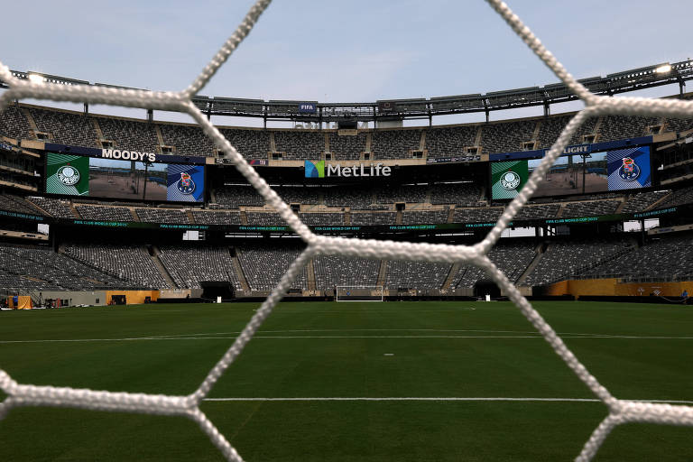 A imagem mostra a visão do campo de futebol através da rede de um gol. No fundo, é possível ver as arquibancadas do Estádio MetLife, com assentos vazios. Há telões visíveis nas laterais, exibindo os logotipos de 'Moody's' e 'Real Madrid'. O campo está bem cuidado e a atmosfera é de um dia claro.