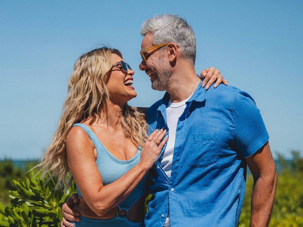 Otaviano Costa e a esposa Flávia Alessandra *** Local Caption *** Um casal está posando para a foto em um ambiente ao ar livre, com um céu azul claro ao fundo. A mulher, com cabelo longo e ondulado, usa um vestido azul claro e óculos de sol. O homem, com cabelo grisalho, veste uma camisa azul e também usa óculos de sol. Eles estão sorrindo um para o outro, demonstrando afeto, enquanto a mulher coloca a mão no peito do homem. Ao redor, há vegetação verde e uma estrutura de madeira ao fundo.