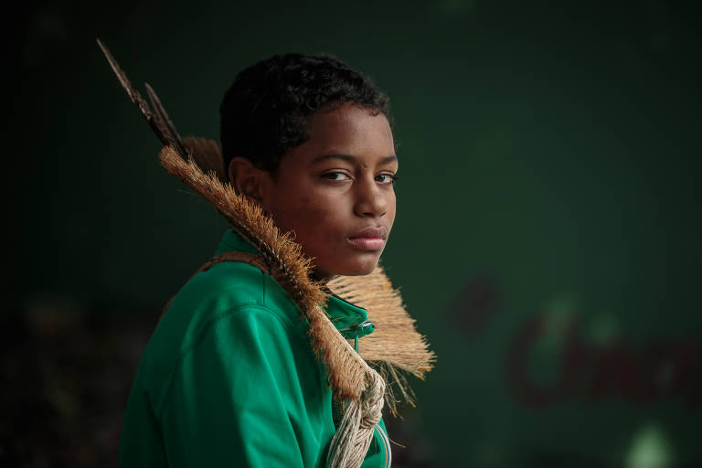 Um jovem com cabelo curto e encaracolado está posando para a foto. Ele usa uma camisa verde e tem uma expressão séria. Nas costas, ele carrega um cocar feito de palha. O fundo é de cor verde escura, criando um contraste com a roupa do jovem.
