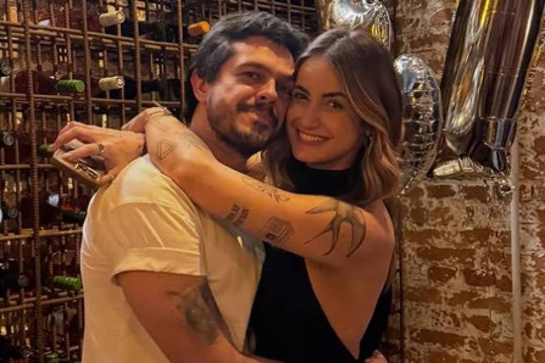 Um casal se abraça em um ambiente decorado com prateleiras de vinho ao fundo. Eles estão sorrindo e parecem felizes. O homem usa uma camiseta branca e a mulher está vestida com um vestido preto. Ao lado deles, há balões em forma do número 17, indicando uma celebração. No chão, há uma sacola da marca Hering.