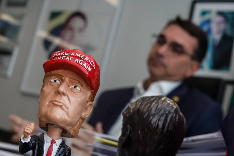 A imagem mostra um boneco com a cabeça de um homem, vestindo um terno e uma gravata, e um chapéu vermelho com a frase "MAKE AMERICA GREAT AGAIN". Ao fundo, há uma pessoa desfocada, aparentemente em uma mesa de escritório, com algumas obras de arte nas paredes. O ambiente parece ser um escritório moderno.
