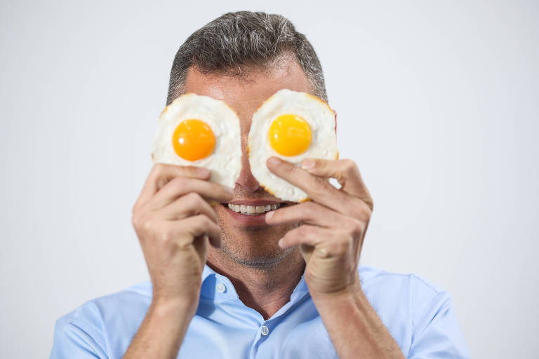 Um homem sorridente está segurando duas fatias de pão com ovos fritos, usando-os como se fossem olhos. Ele está vestindo uma camisa azul e tem cabelo curto e grisalho. O fundo da imagem é claro e neutro, destacando a expressão divertida do homem.