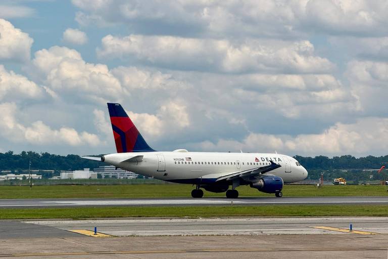 Um avião da Delta Airlines está taxiando na pista de um aeroporto. O avião é de cor branca com detalhes em azul e vermelho, e possui o logotipo da Delta na fuselagem. O céu está parcialmente nublado, com nuvens brancas e um fundo azul. Ao fundo, é possível ver uma área verde e algumas construções.