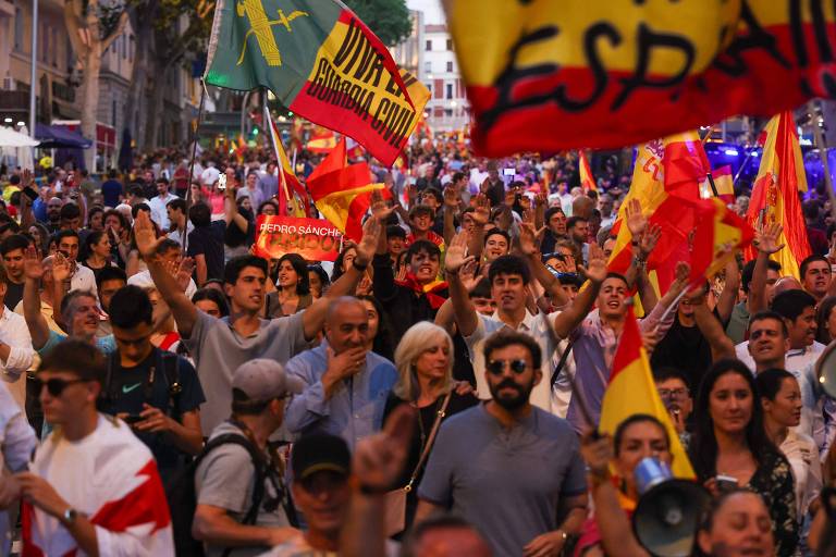 The image shows a large crowd at a demonstration, with people raising Spanish flags. Some people are smiling and waving, while others hold signs. The atmosphere is festive, with many people wearing t-shirts and clothing in colors representing the Spanish flag. In the background, there are buildings and a celebratory atmosphere.