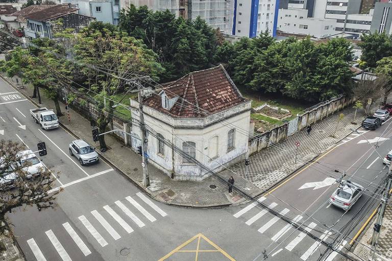 A imagem mostra uma esquina com uma casa antiga de paredes brancas e telhado de duas águas. A casa está cercada por um pequeno jardim e árvores ao fundo. Na rua, há veículos estacionados e em movimento, além de faixas de pedestres visíveis. O ambiente urbano é caracterizado por prédios modernos ao fundo, contrastando com a arquitetura da casa.